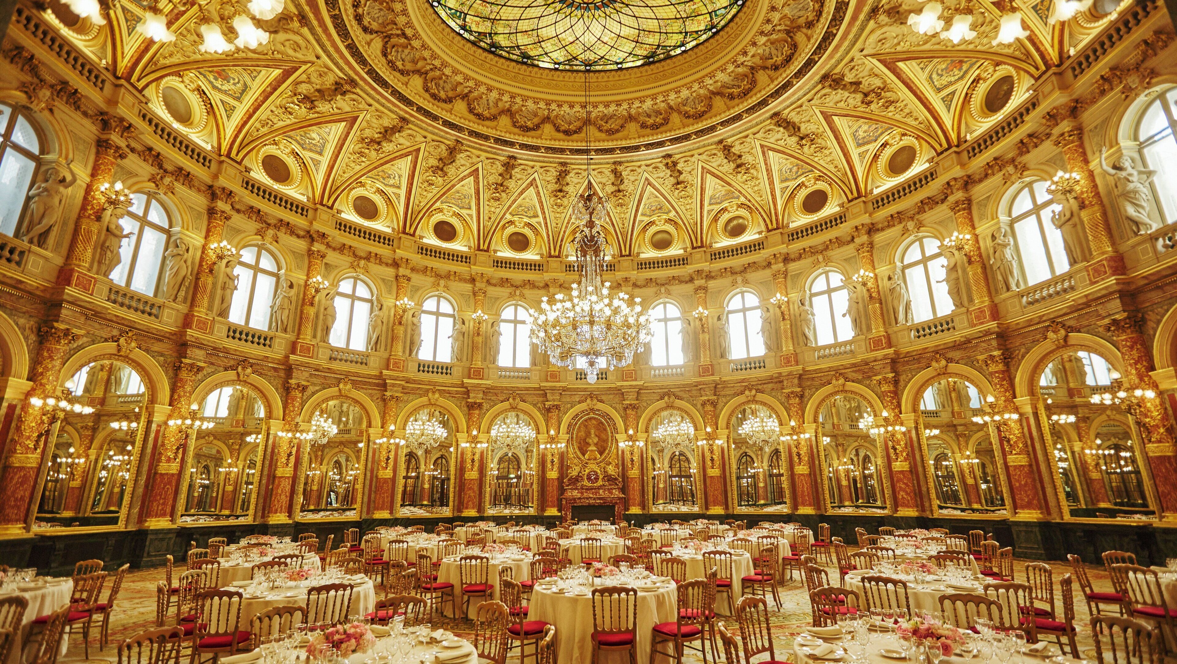 Un salón imponente con techo dorado ornamentado, candelabros y varias mesas redondas dispuestas para comer.