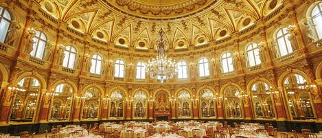 Un salón imponente con techo dorado ornamentado, candelabros y varias mesas redondas dispuestas para comer.
