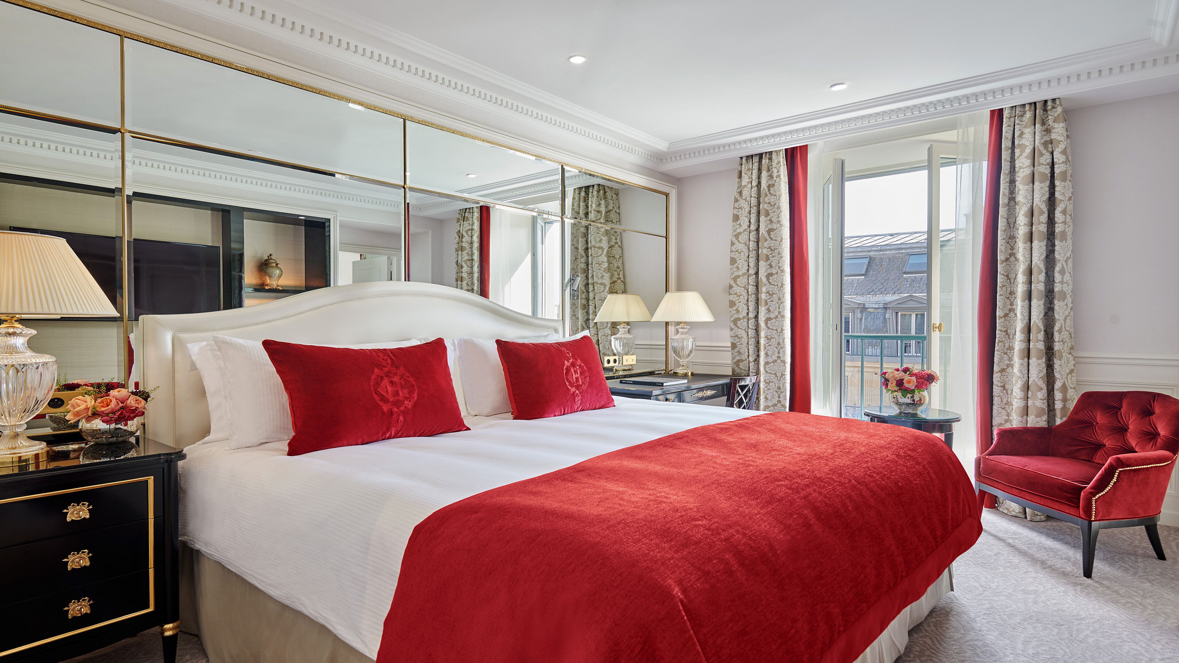 Une chambre d’hôtel avec un grand lit, un fauteuil rouge, une table de chevet avec une lampe et une vue sur la ville par la fenêtre.