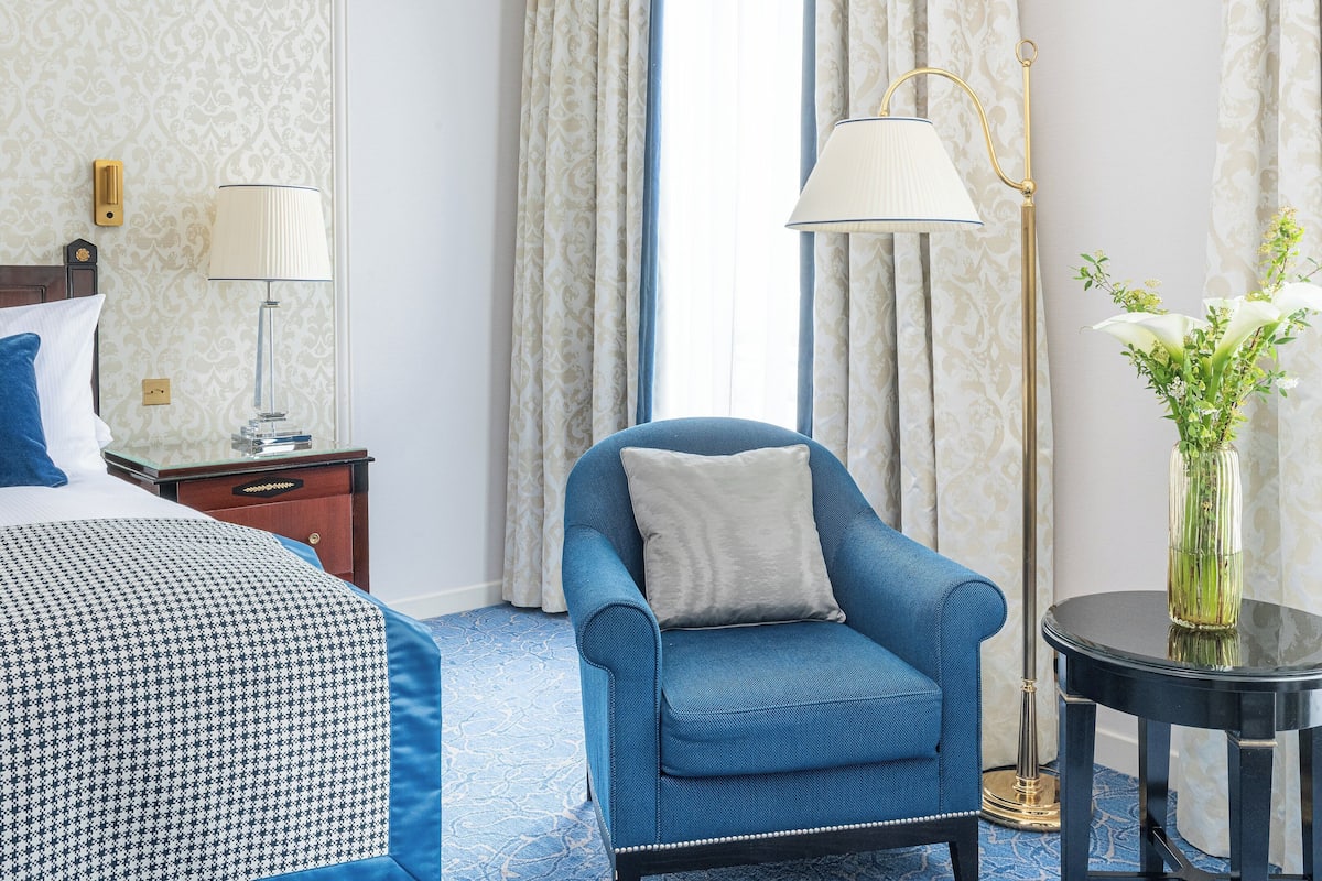 A hotel room with a bed, a blue armchair, a nightstand, a lamp, and a vase with flowers.