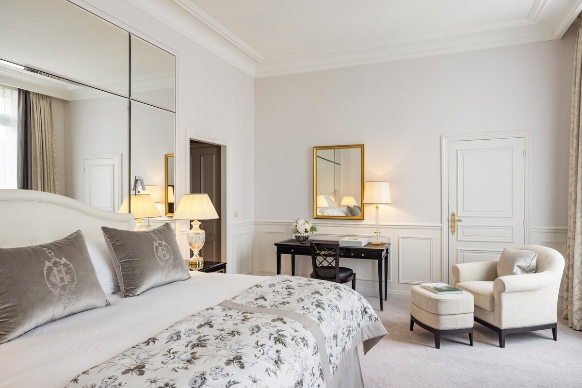 A hotel room with a large bed, a desk, a chair, and a small ottoman.