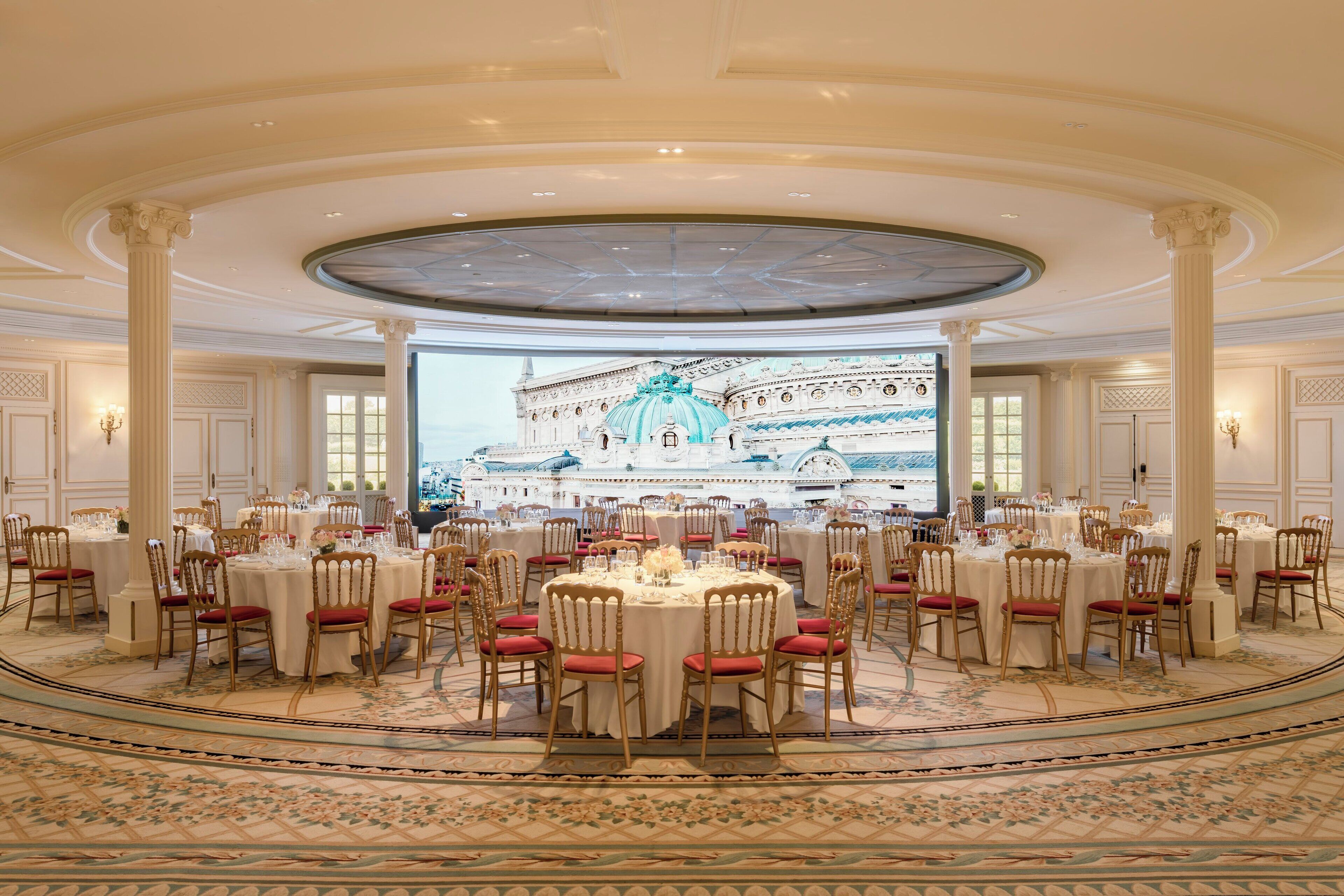 A grand ballroom with round tables, red upholstered chairs, and a large projection screen displaying a building.