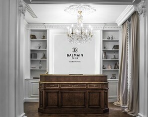 A Balmain Paris hair salon with a classic design, featuring a wooden reception desk, ornate chandeliers, and elegant shelving with decorative items.