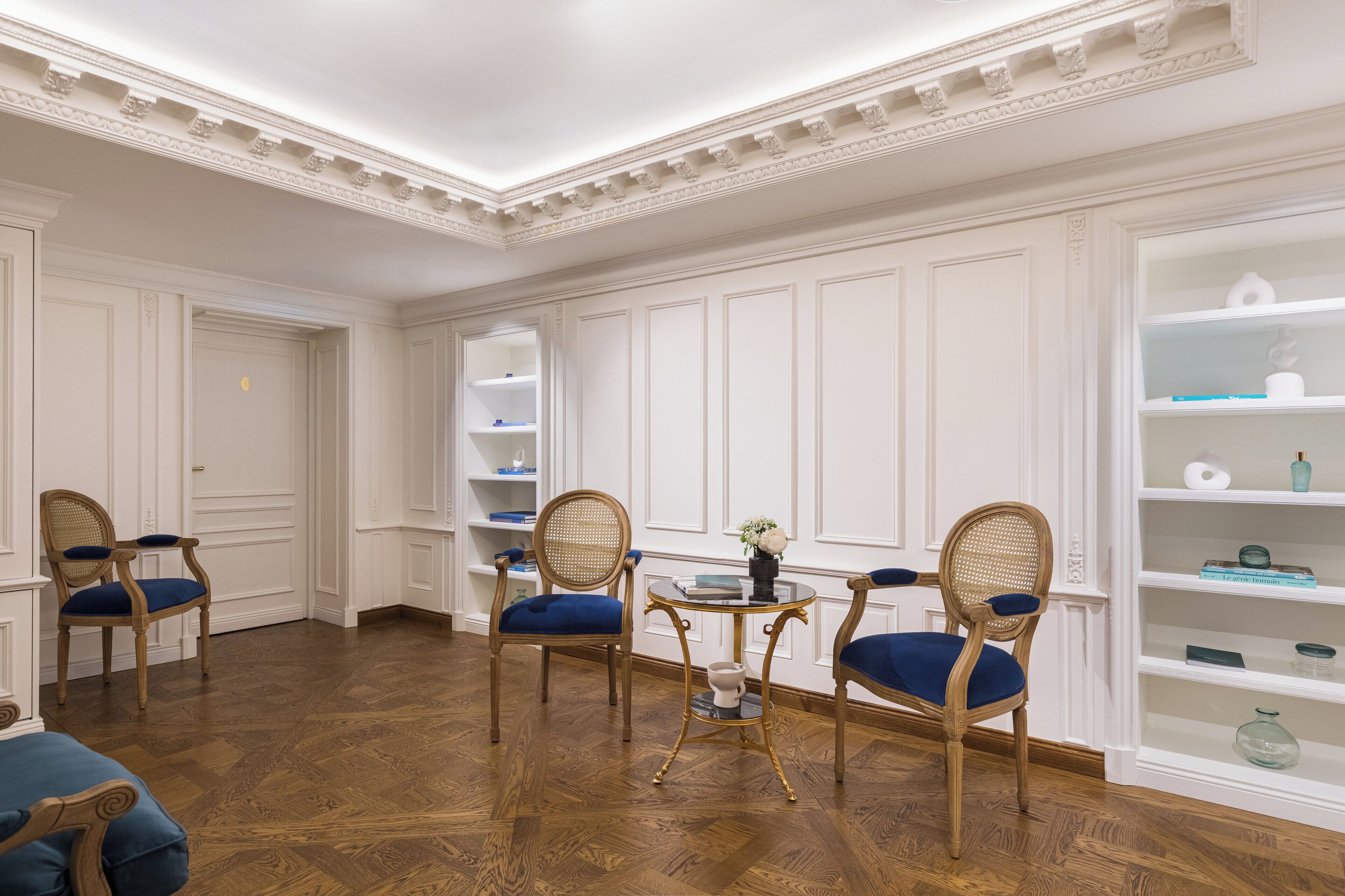 Una habitación con suelo de madera, paredes blancas con paneles y dos sillones tapizados en azul con estructura de madera.