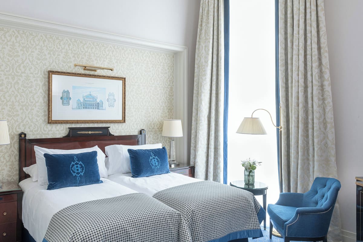 A hotel room with a large bed, two pillows, a nightstand, a lamp, a chair, and a framed picture on the wall.