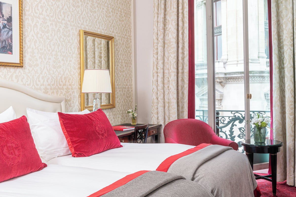 A hotel room with a large bed, a desk, a chair, a lamp, and a view of the city through the window.