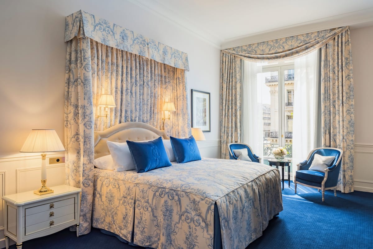 A hotel room with a large bed, two bedside lamps, a small table with a vase, a chair, and a view of the city through large windows.
