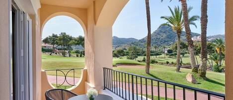 A balcony with a table and chairs, overlooking a golf course with palm trees and a mountain in the background.