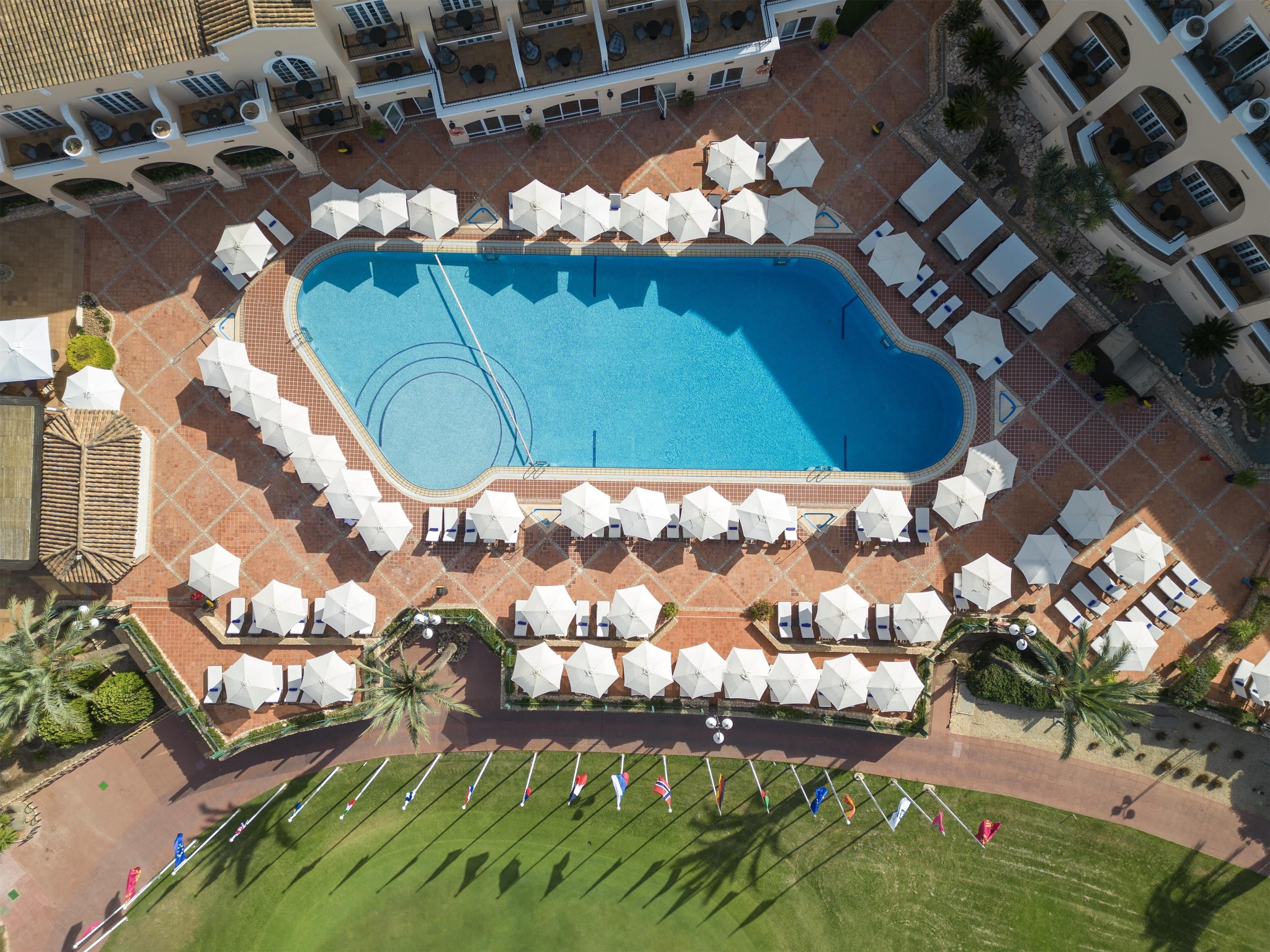 En vy från ovan av ett hotell med en central pool omgiven av solstolar och parasoller, en gräsmatta kantad av flaggor och en plattbelagd uteplats.