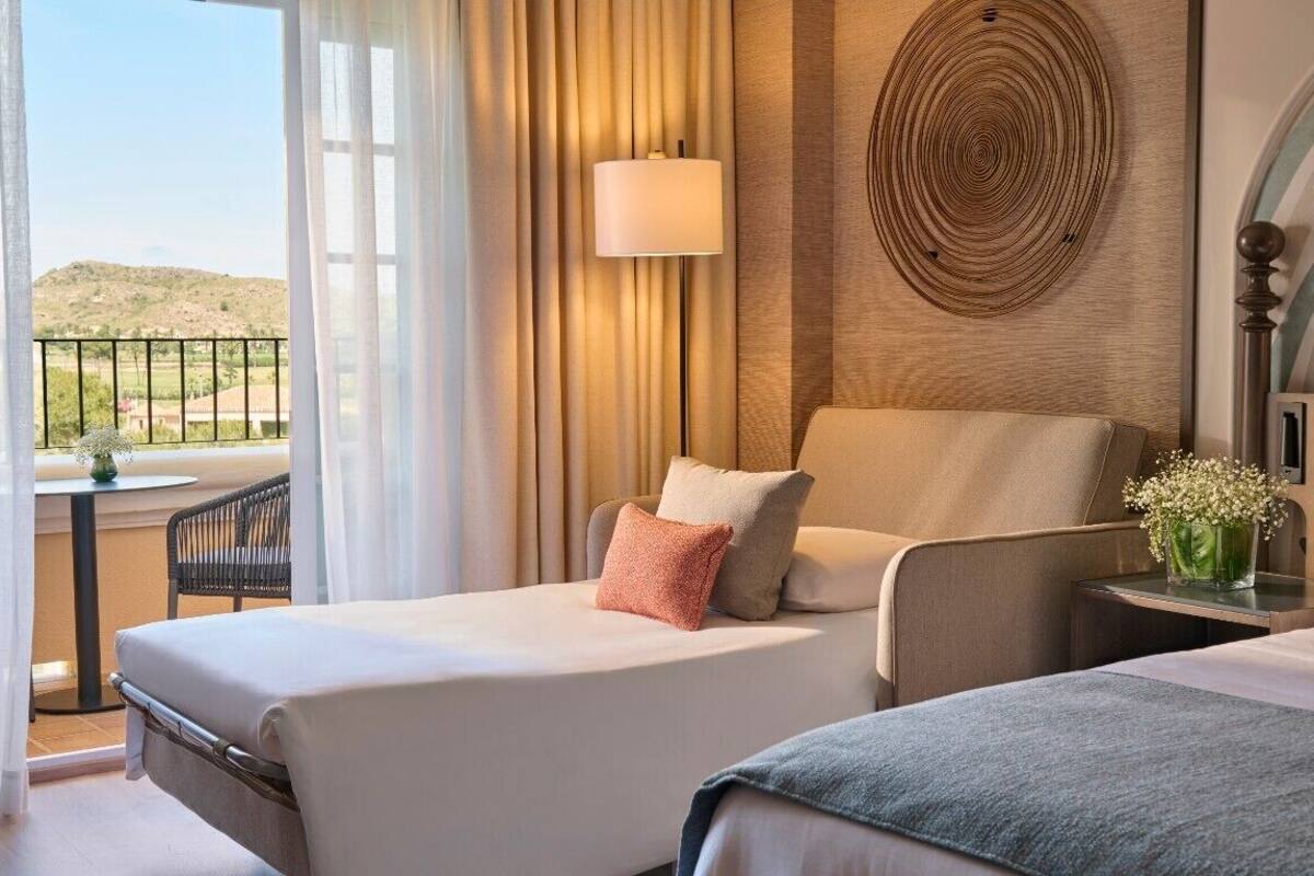 Une chambre d’hôtel avec deux lits, un balcon doté d’une table et d’une chaise, une lampe et un élément décoratif mural.