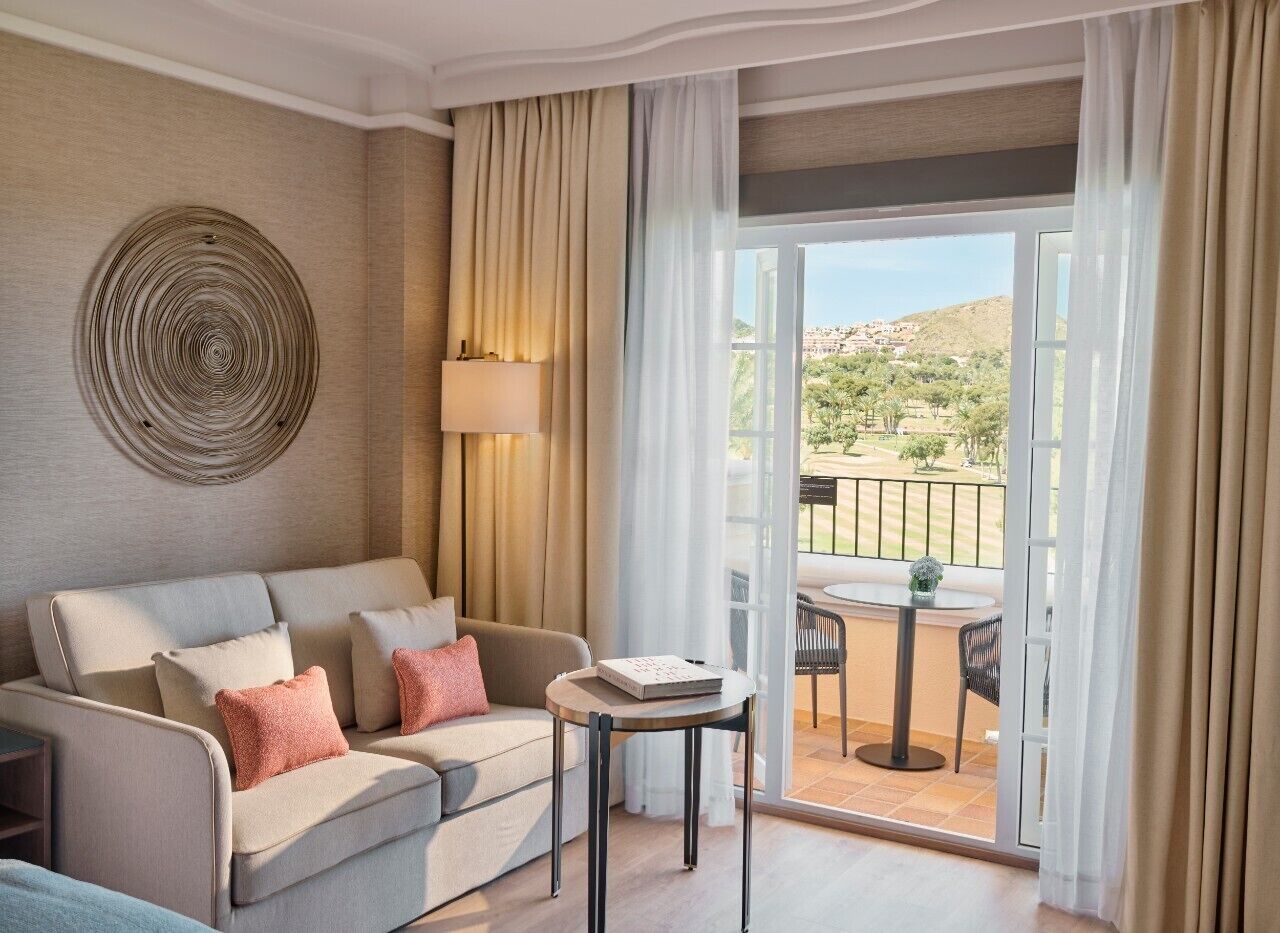 Quarto de hotel com sofá, uma mesinha e vista para uma varanda com mobiliário externo.