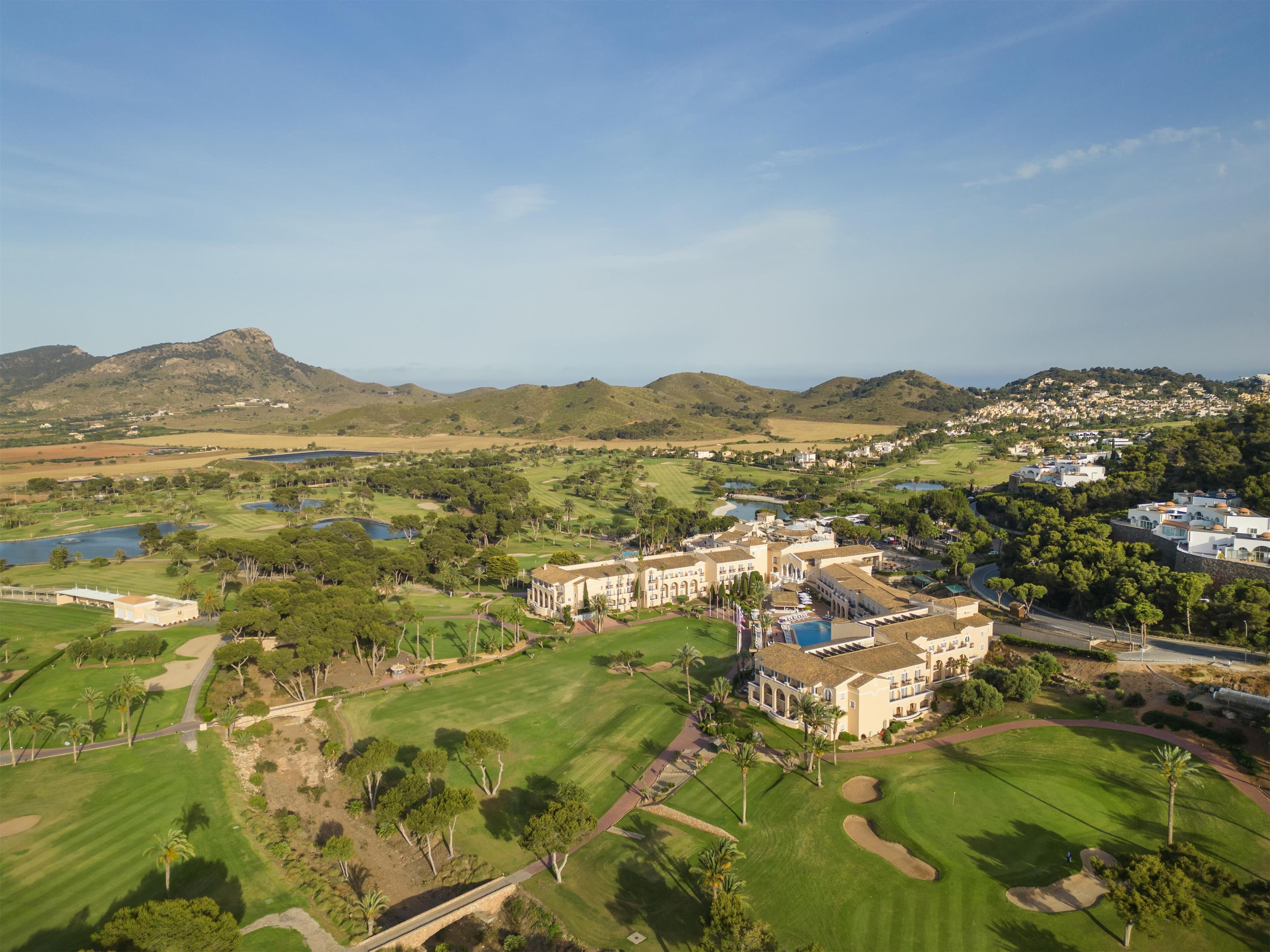 A golf course with a large building in the foreground, surrounded by lush greenery and hills in the background.