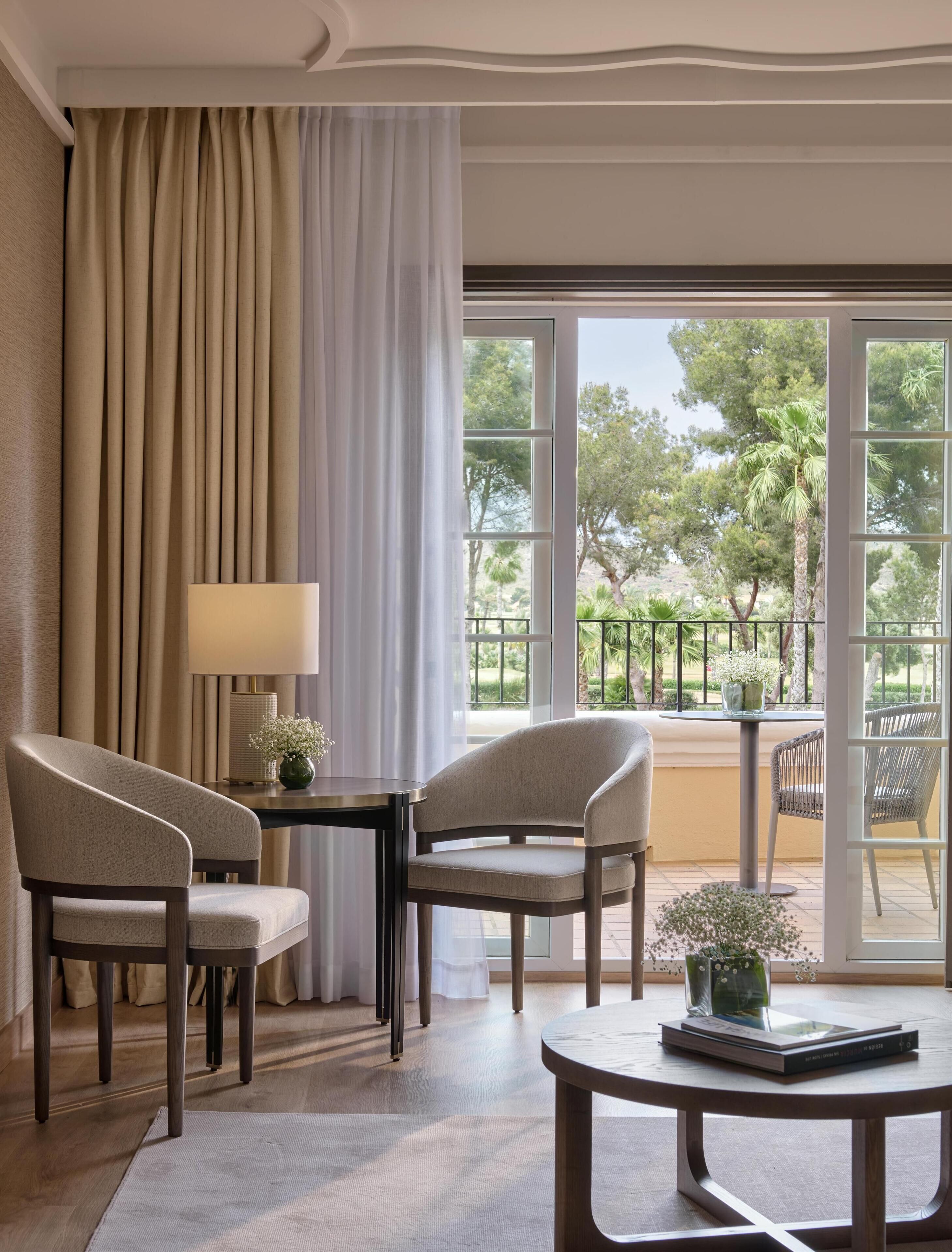 Une pièce avec deux chaises, une table et une lampe. Il y a une grande fenêtre donnant sur des arbres et un balcon.