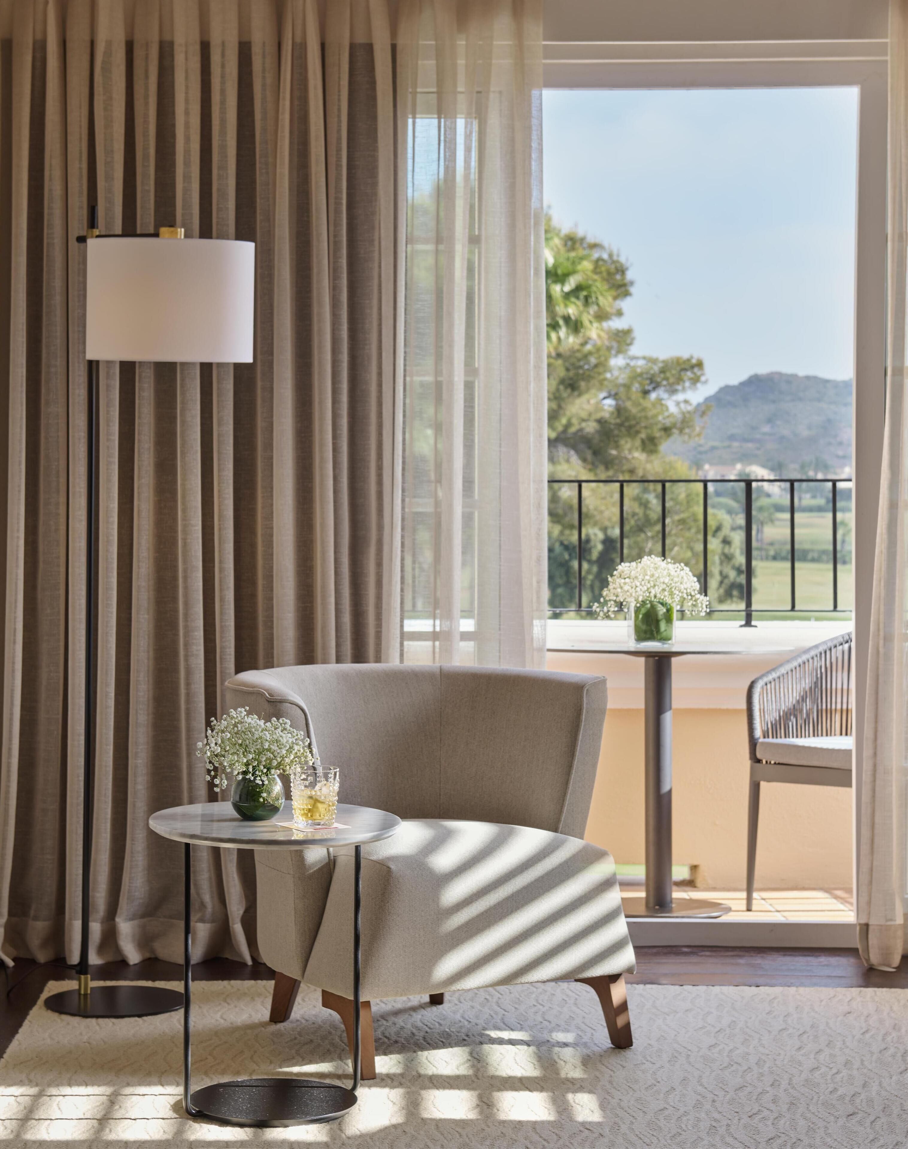 A room with a large window, a chair, a small table with a vase of flowers, and a lamp.
