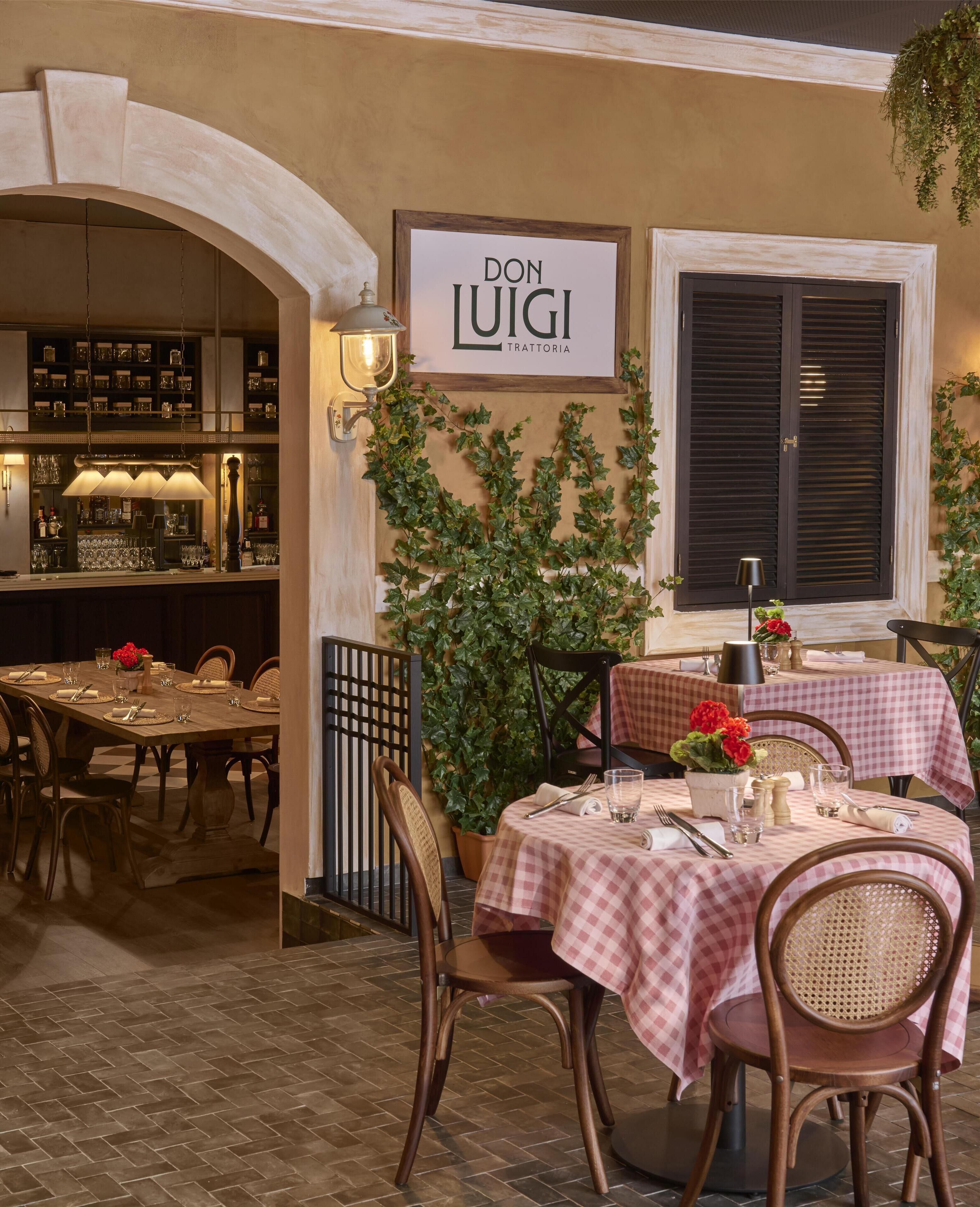 An outdoor dining area with tables set for two, a bar area with shelves of bottles, and a sign reading 'DON LUIGI TRATTORIA'.