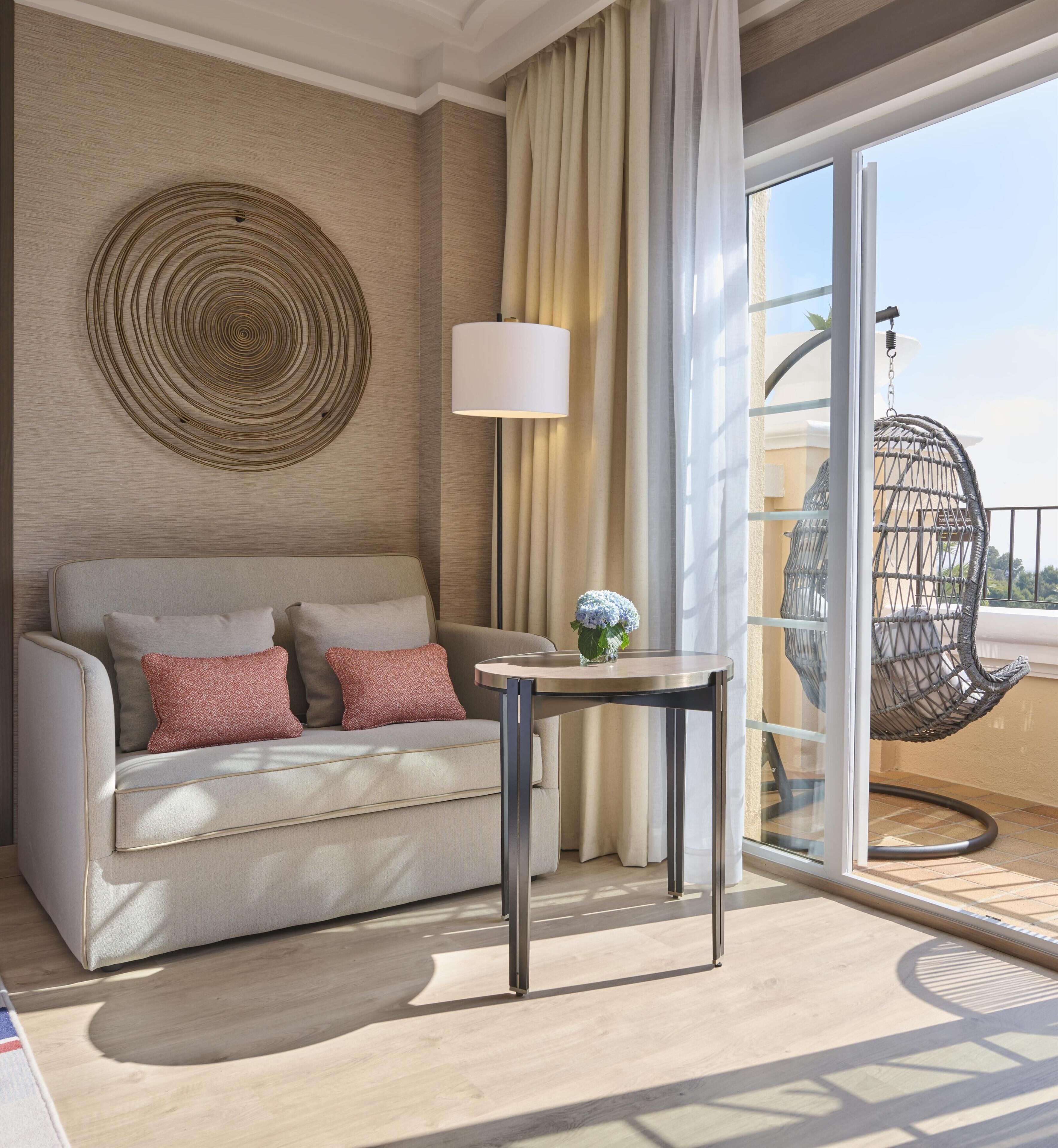 Quarto de hotel moderno com sofá, mesa, luminária e vista para a varanda.