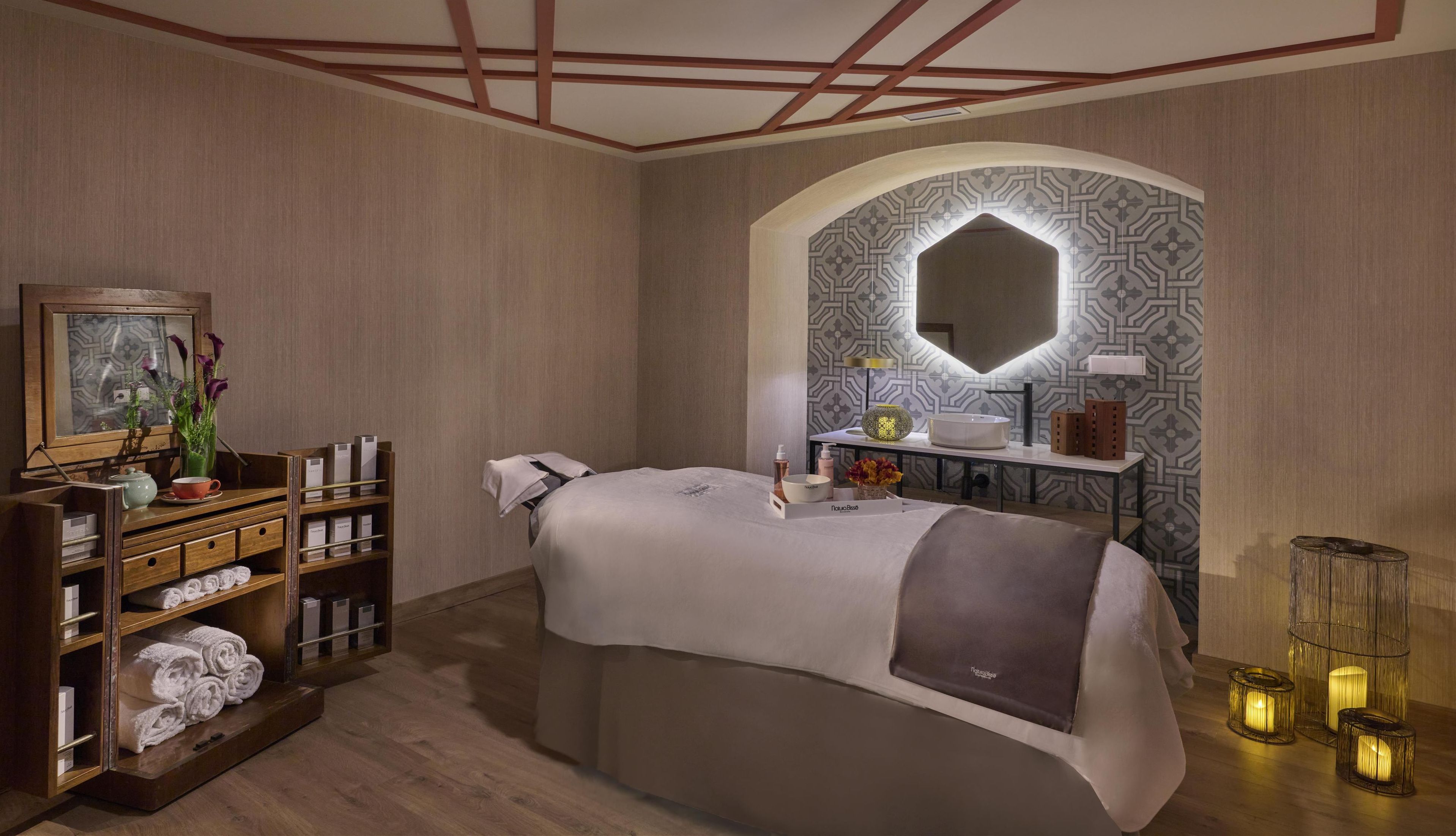 Un moderno spa con una cama, un tocador con espejo, una estantería con toallas y una pared con diseño geométrico.