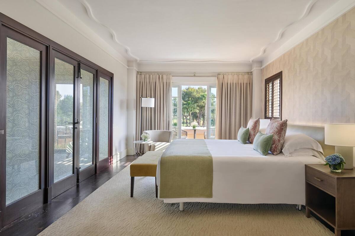 Une chambre d’hôtel avec un grand lit, une table de chevet avec une lampe, une petite table d’appoint et une vue sur l’extérieur grâce à une porte-fenêtre coulissante.