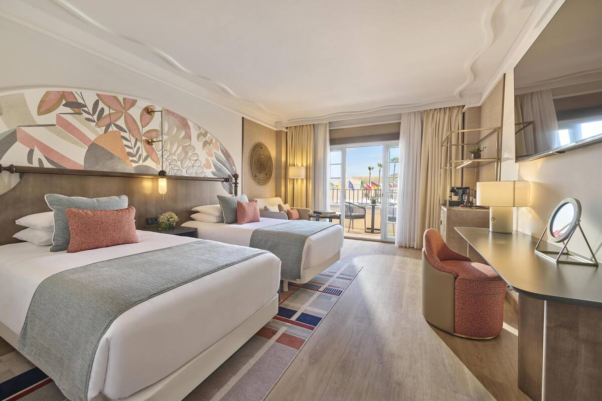Une chambre d’hôtel avec deux lits, un bureau, une chaise, une grande fenêtre et un mur décoratif.