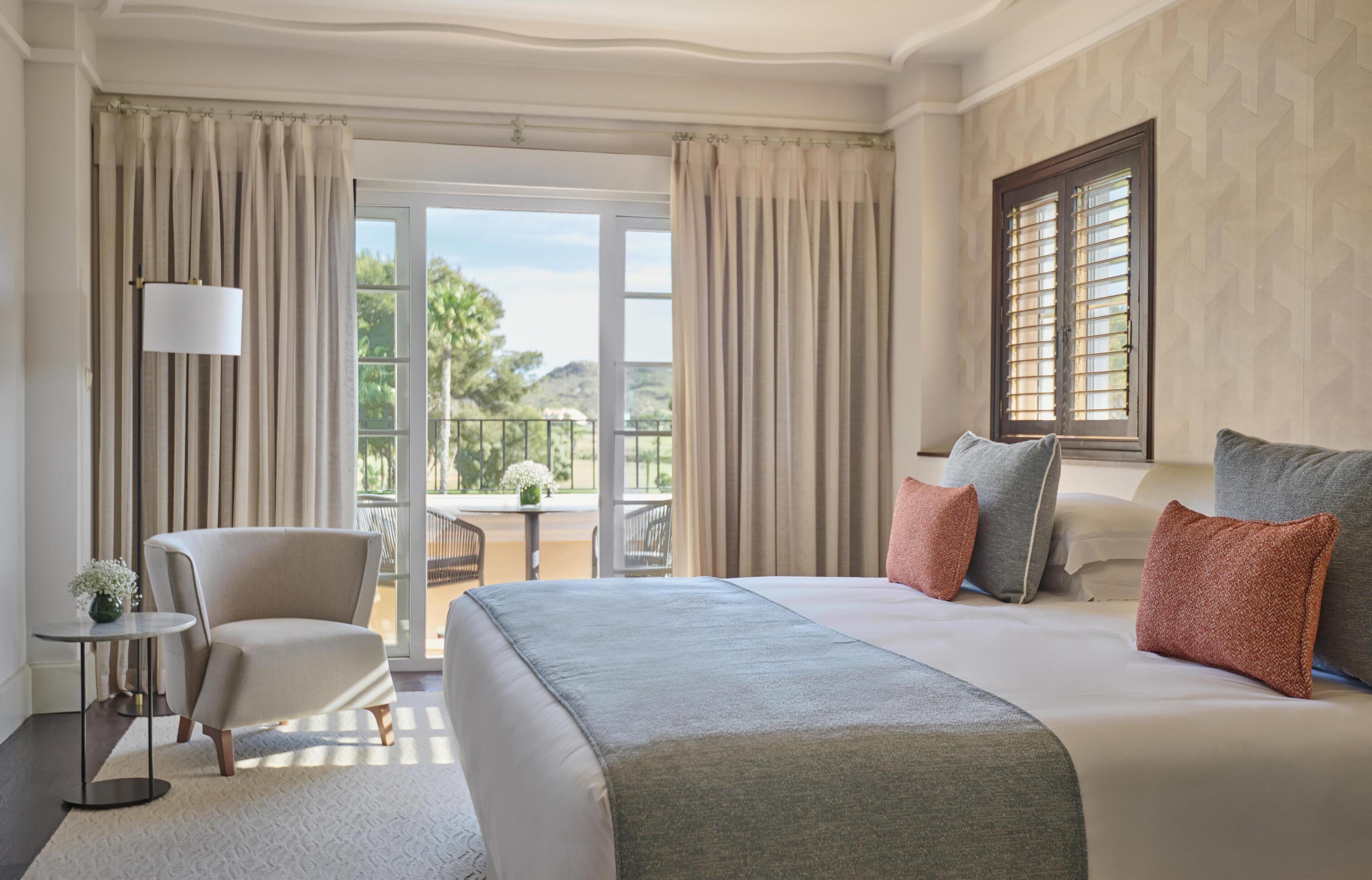 Habitación de hotel con una cama grande, una silla, una mesita y un balcón con vistas a zonas verdes.