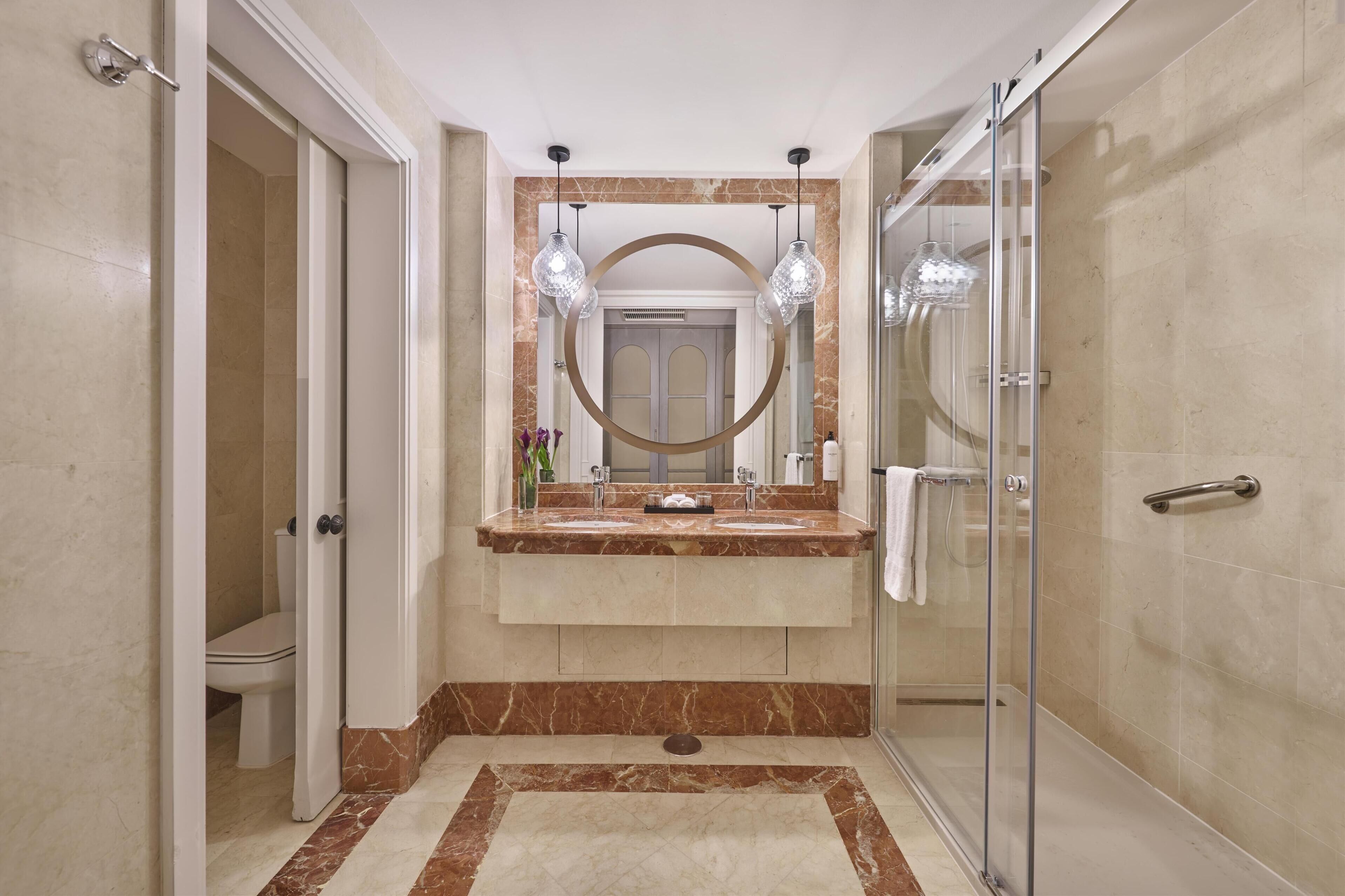 Un baño moderno con cabina de ducha de vidrio, tocador de mármol y un espejo grande con marco circular.