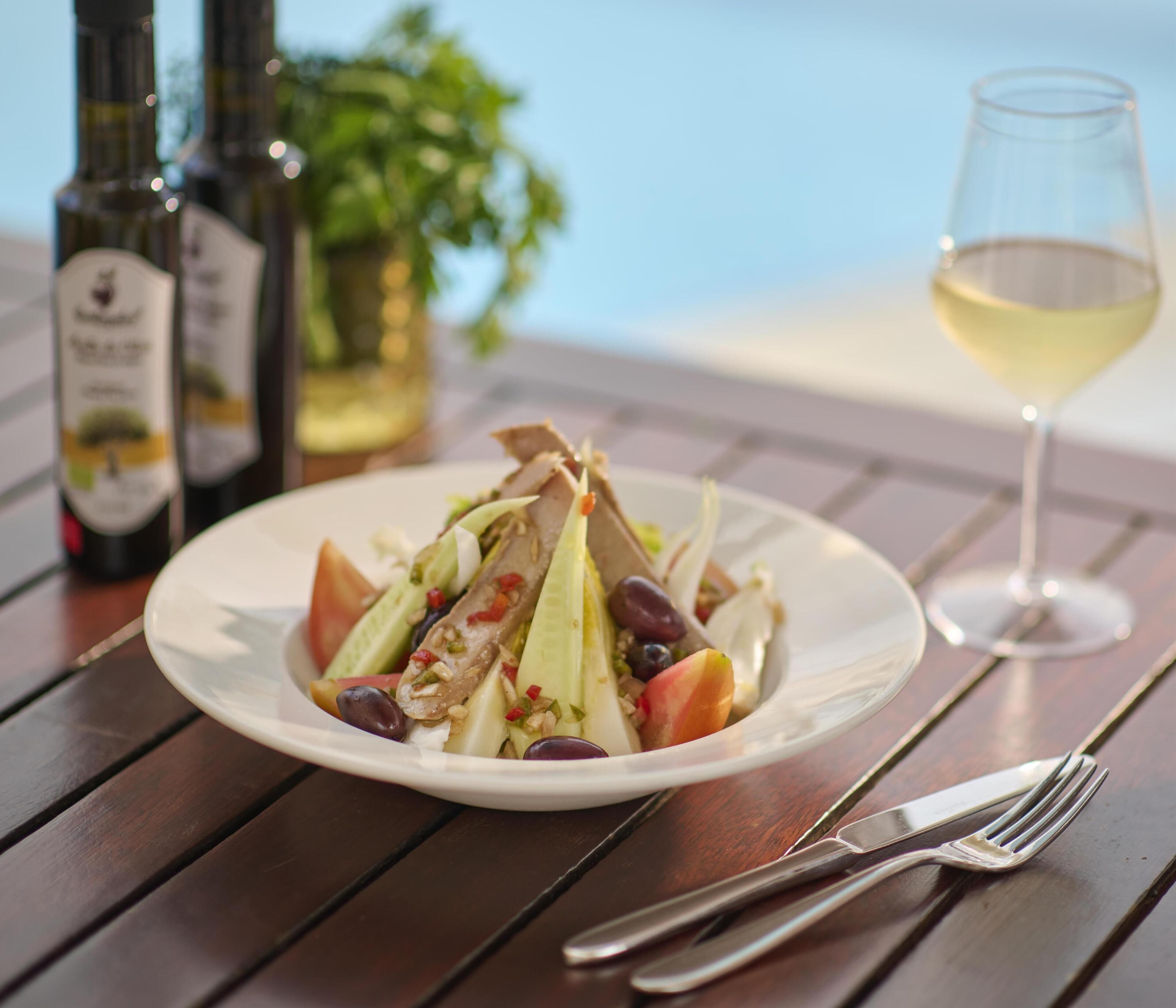 Ensalada en un plato con una copa de vino blanco y botellas de vino sobre una mesa de madera.
