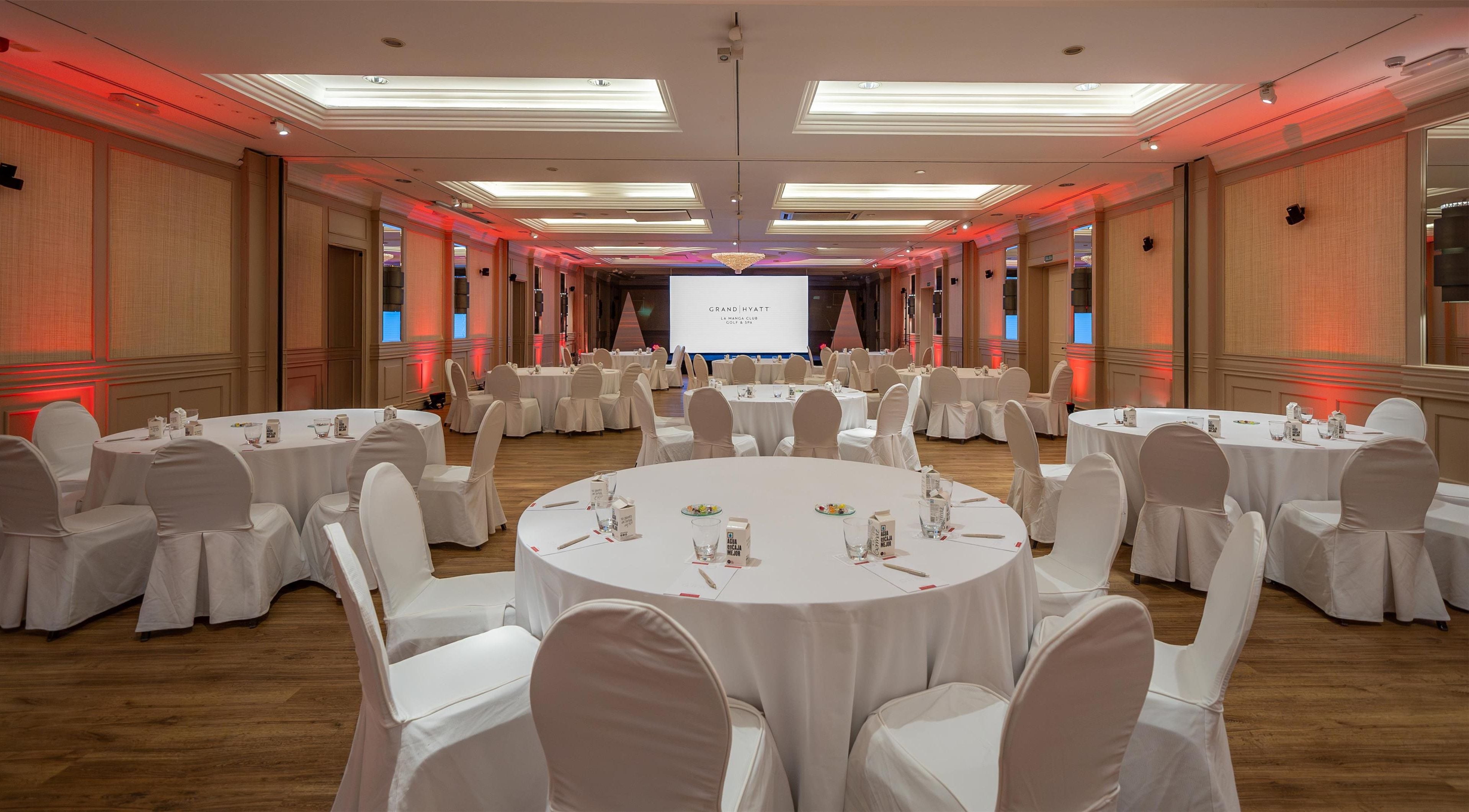 A large, well-lit conference room with round tables and white chairs, equipped with nameplates and water glasses.