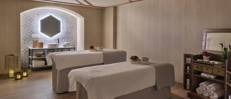 Un cuarto de spa con dos camillas de masaje, un tocador con espejo y un estante con toallas.