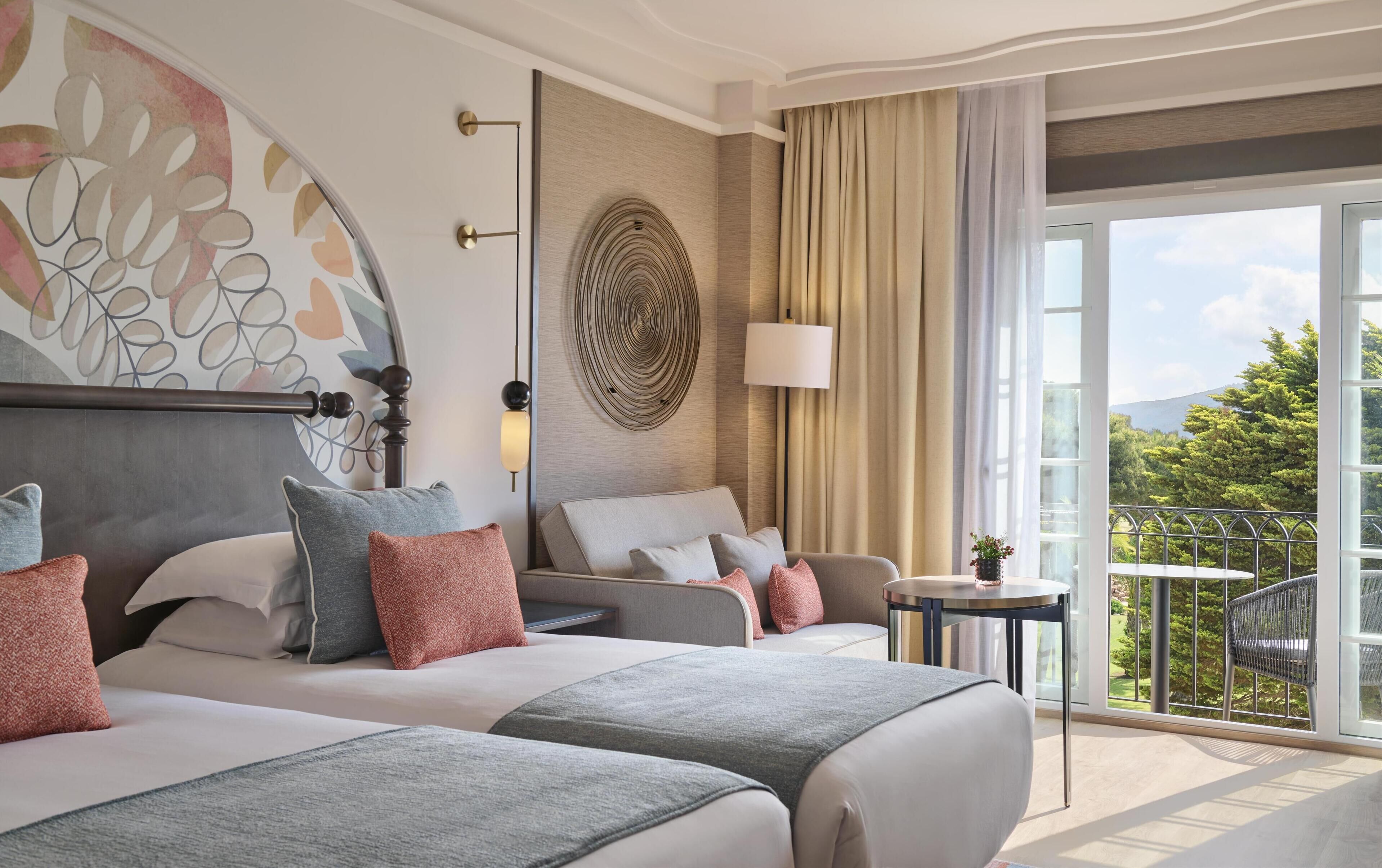 Habitación de hotel con dos camas, un ventanal con vistas a zonas verdes, un adorno redondo en la pared y una mesita con una lámpara.