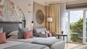 A hotel room with two beds, a large window with a view of greenery, a round decorative wall piece, and a small table with a lamp.