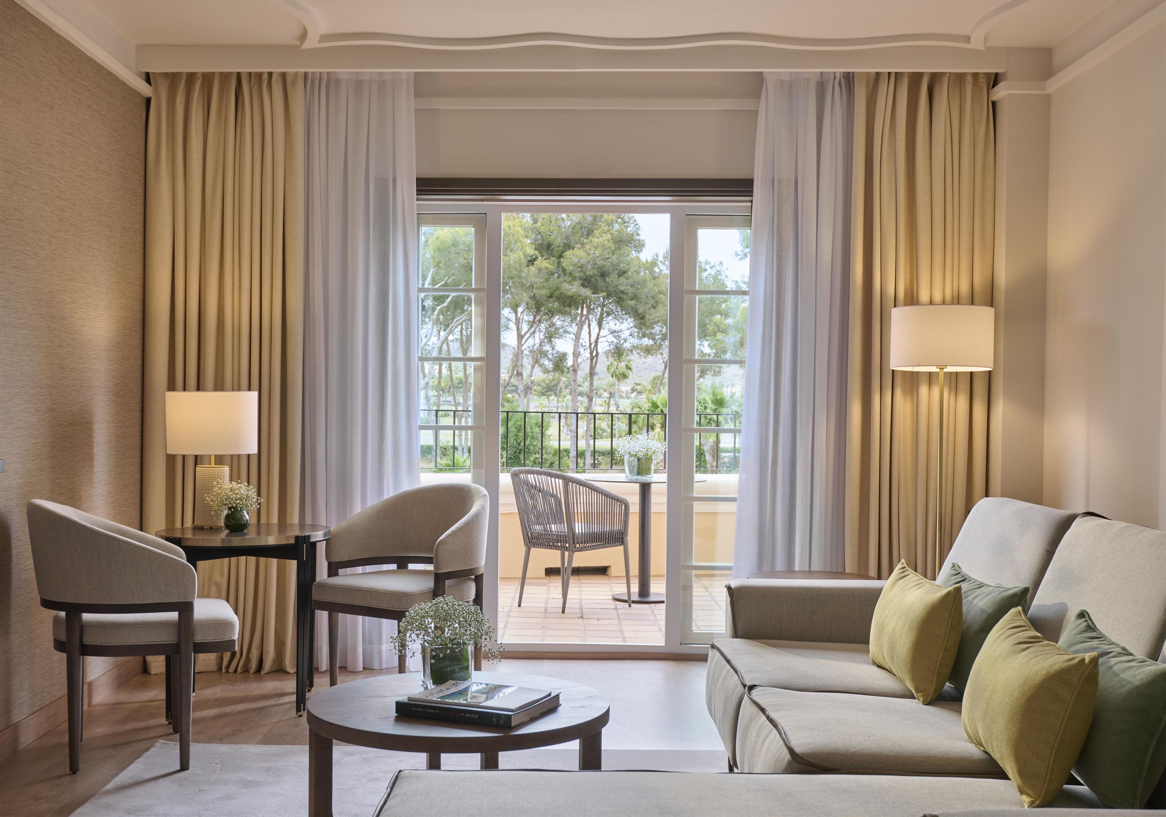 A hotel room with a sofa, two armchairs, a coffee table, a lamp, and a view of the outdoors through large windows.