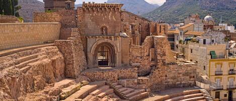 An ancient amphitheater with stone steps and a central stage, surrounded by historic buildings and mountains in the background.
