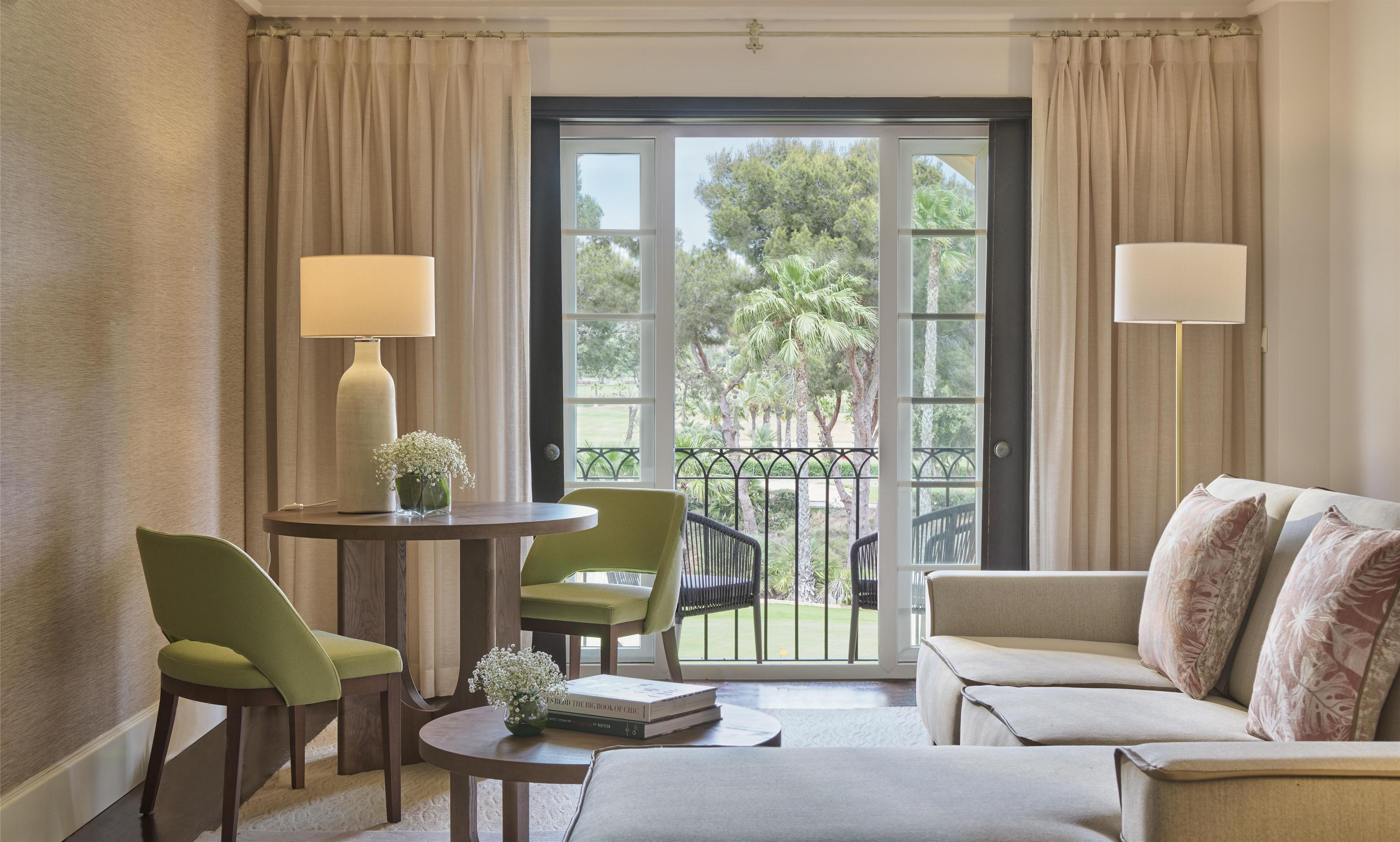 A room with a sofa, two chairs, a small table, a lamp, and a view of greenery through a glass door.