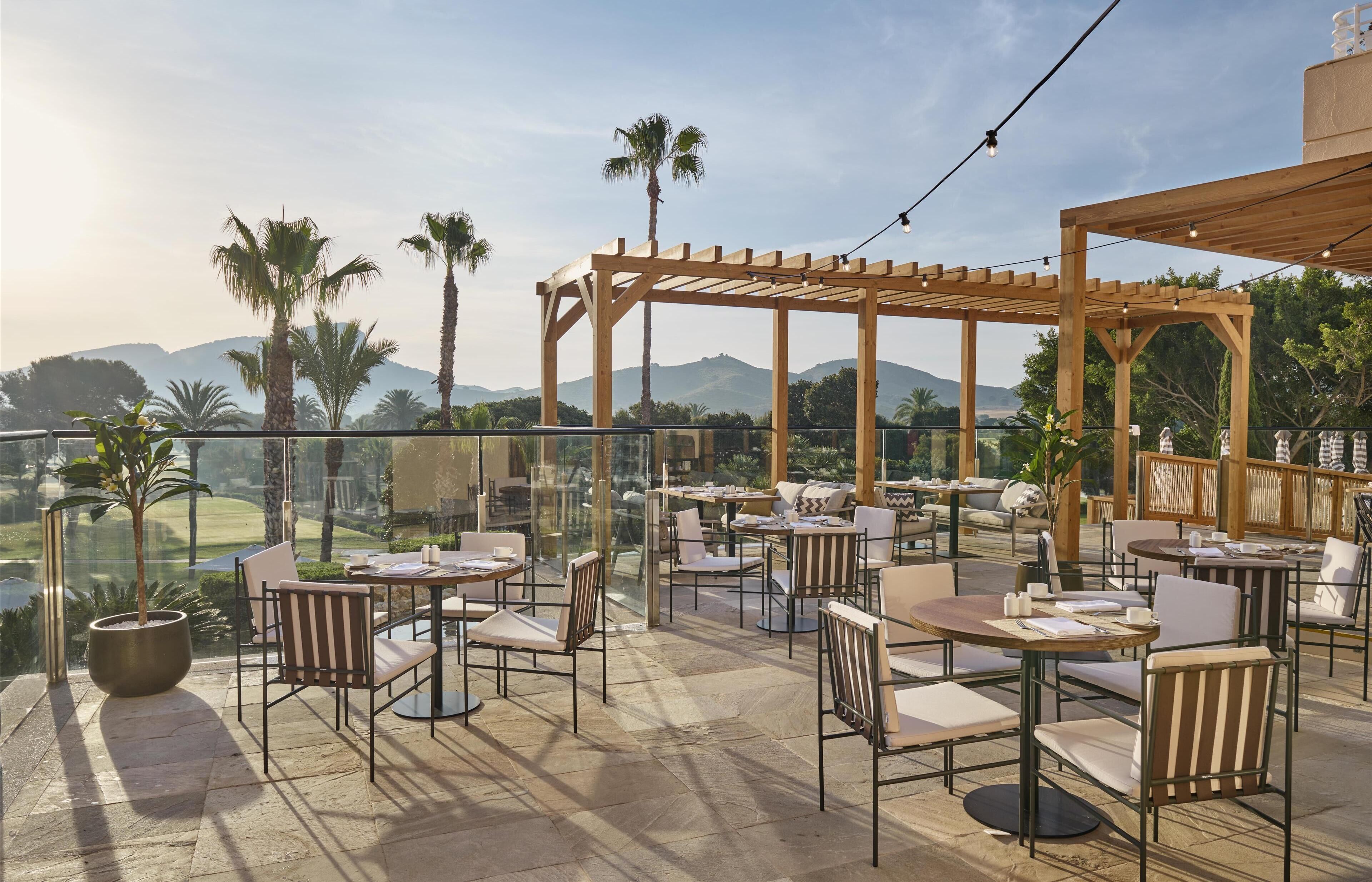 An outdoor patio with tables and chairs, a wooden pergola, and a view of a golf course and mountains.
