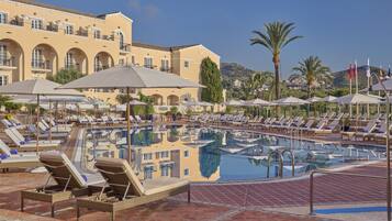 A pool area with lounge chairs and umbrellas, a large building with arches, and palm trees in the background.