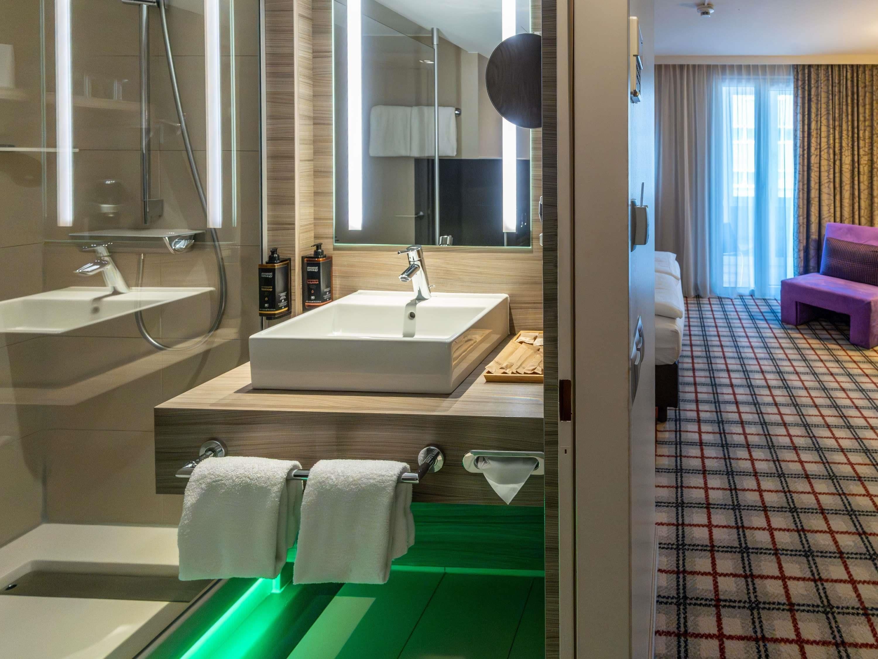 A modern bathroom with a large mirror, two sinks, a shower, and a green-lit tub.