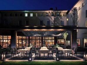 A hotel with an outdoor dining area featuring tables and chairs, a covered patio, and a restaurant visible through large windows.