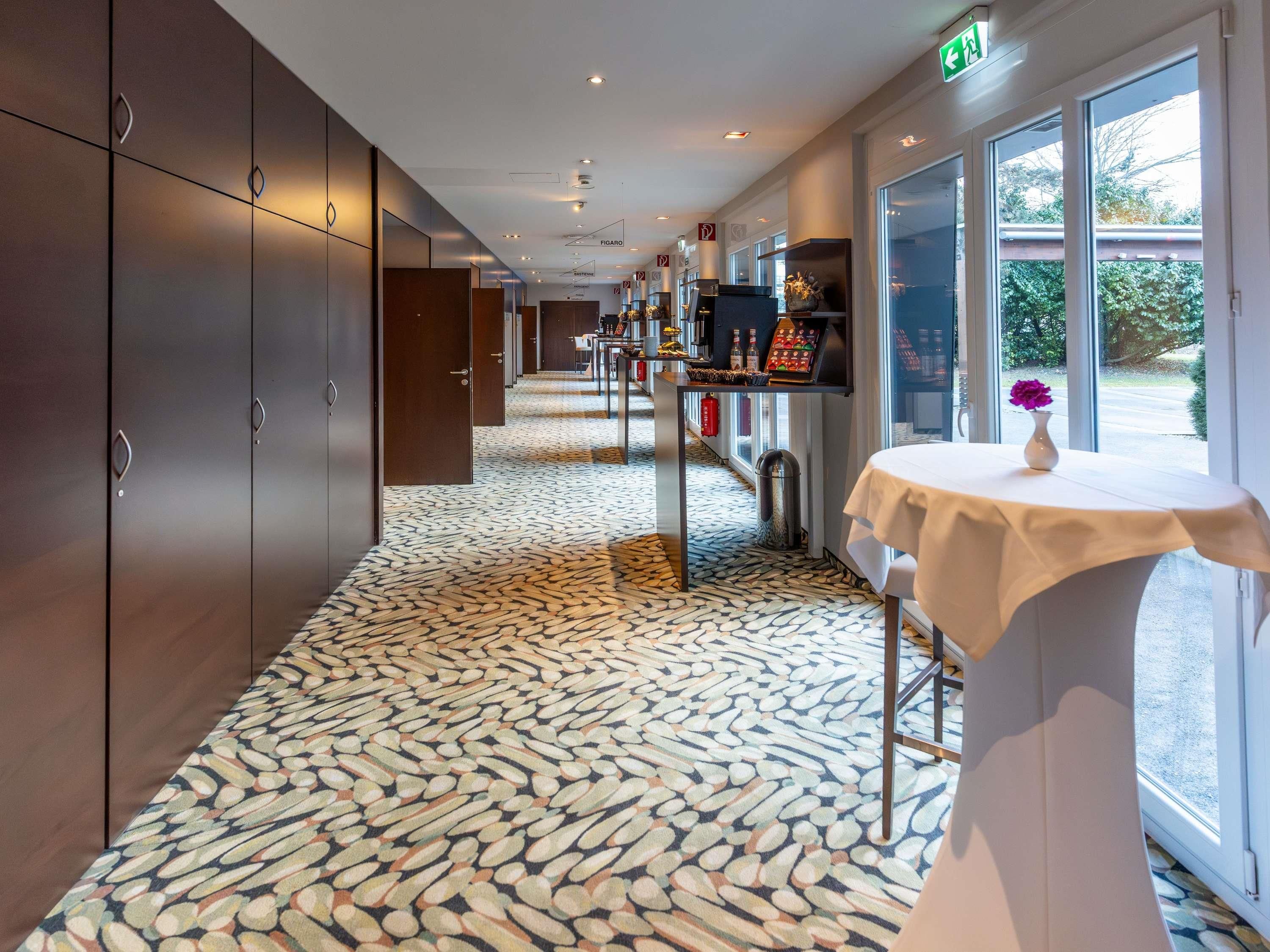 Un couloir d’hôtel avec un sol à motifs, une table sur laquelle est posé un vase, et une vue par la fenêtre.