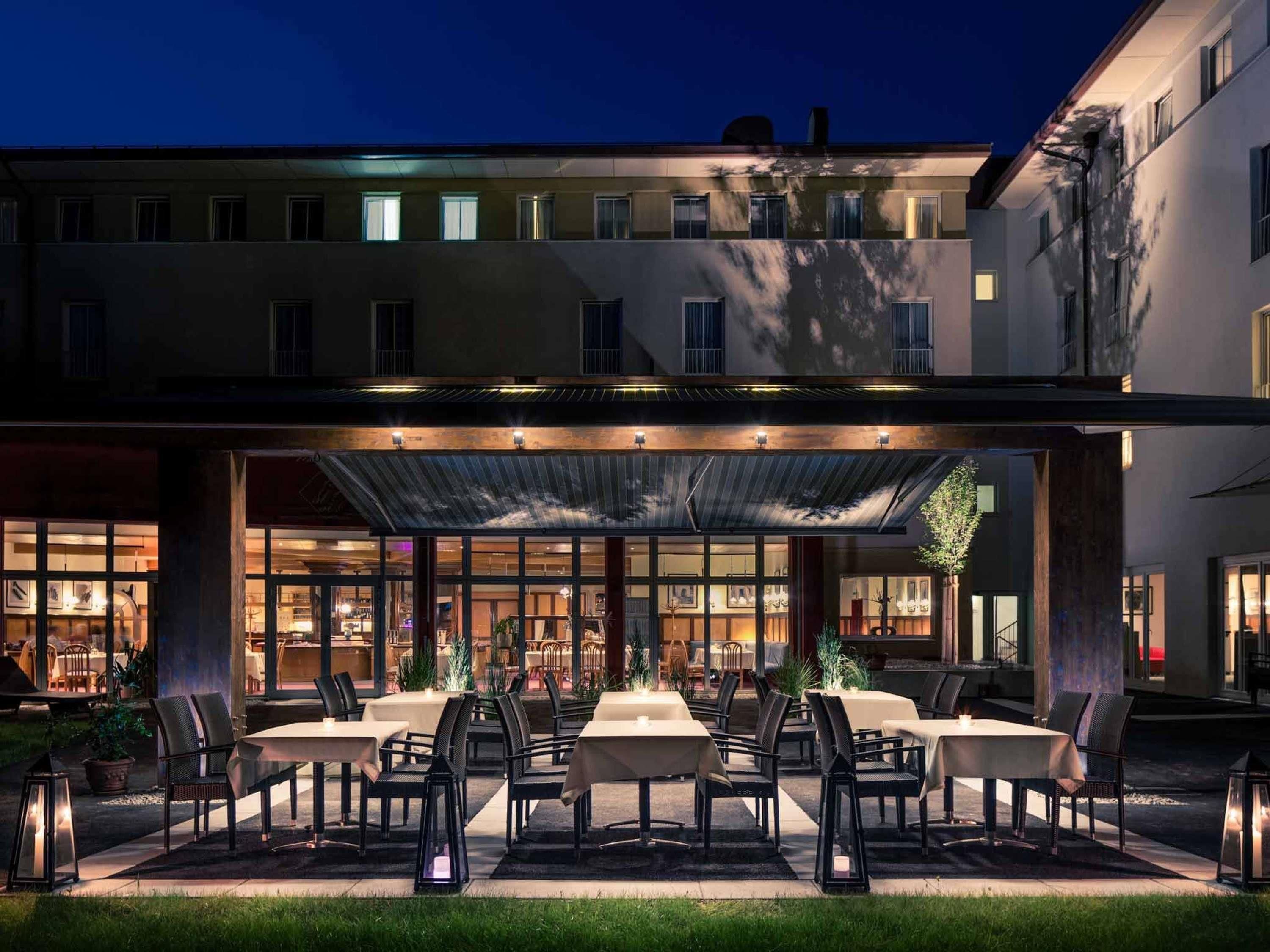 Un espace extérieur abrité, aménagé pour les repas, avec des tables et des chaises, un bâtiment doté de nombreuses fenêtres et un ciel dégagé.