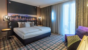 A hotel room with a large bed, a view of a cityscape, a patterned carpet, and a seating area with a table and chairs. - Mercure Salzburg City (Salzburg)