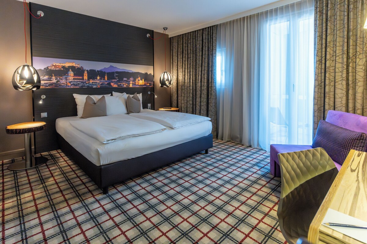 A hotel room with a large bed, a view of a cityscape, a patterned carpet, and a seating area with a table and chairs.