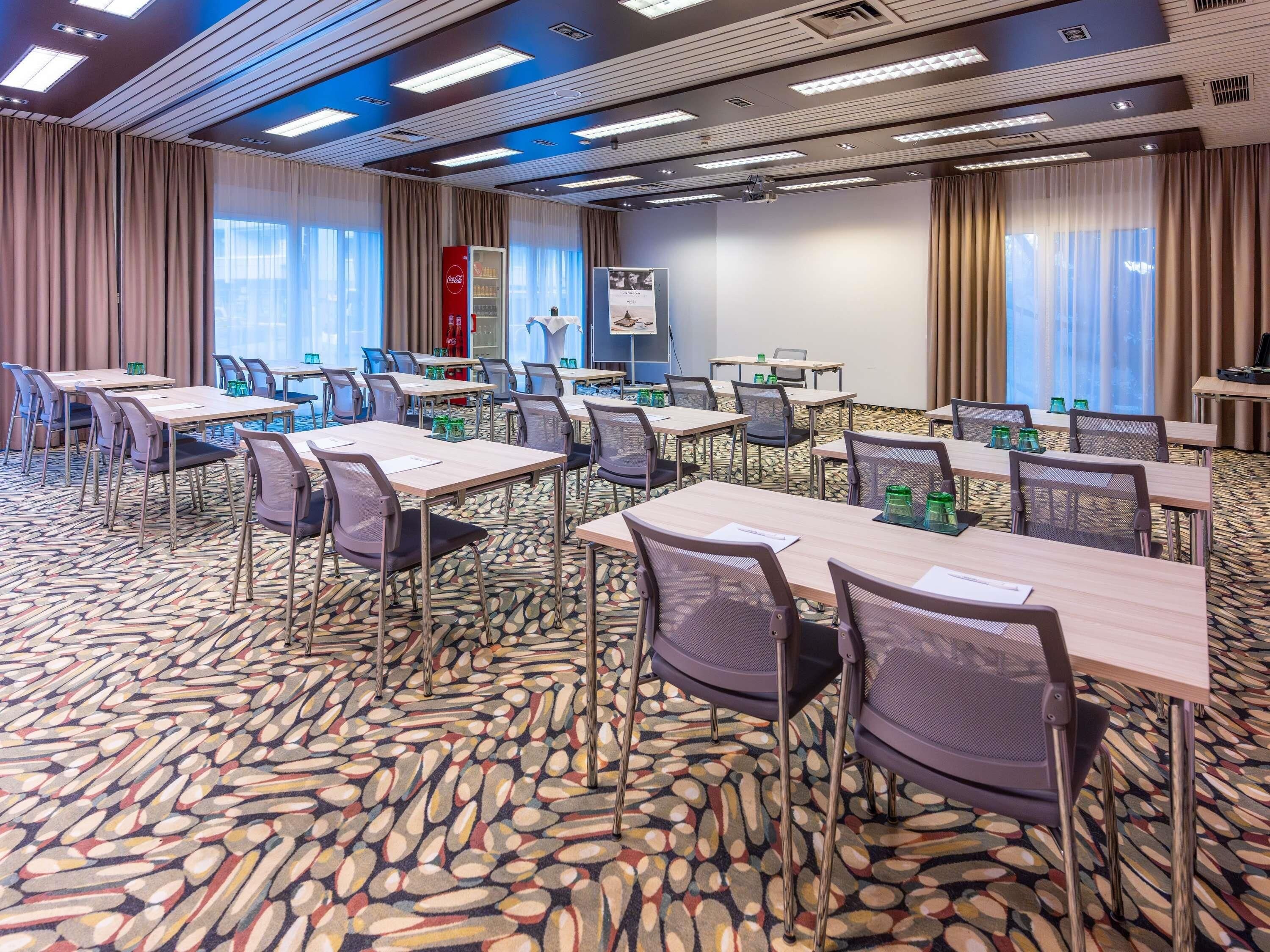 Une salle de conférence équipée de tables, de chaises, d’un écran de projection et d’un réfrigérateur contenant des boissons Coca-Cola.