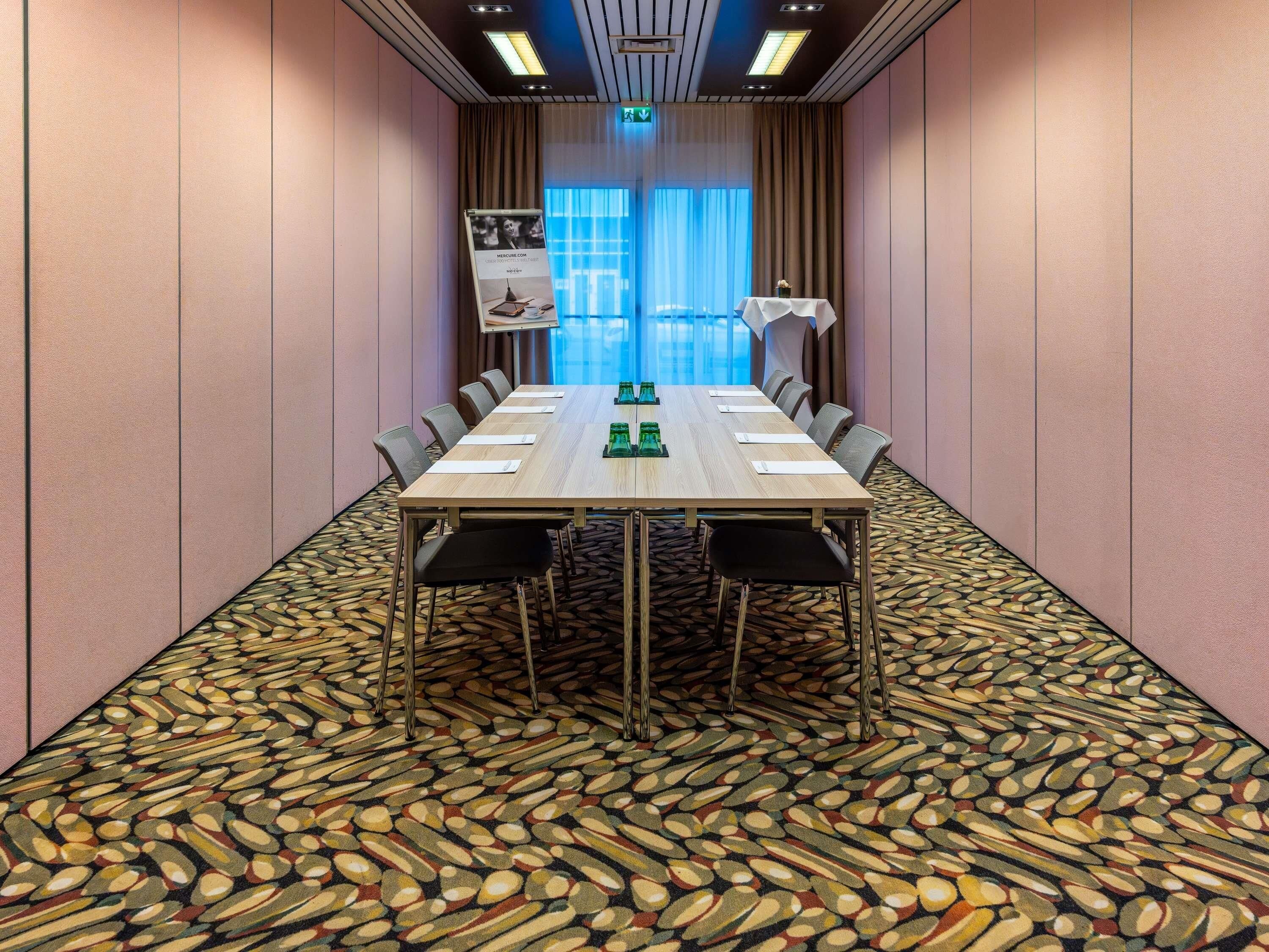 A conference room with a long table, chairs, a patterned carpet, and a view of the outdoors through large windows.