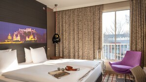 A hotel room with a large bed, a purple chair, a view of a cityscape, and a painting on the wall. - Mercure Salzburg City (Salzburg)