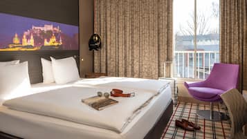 A hotel room with a large bed, a purple chair, a view of a cityscape, and a painting on the wall.