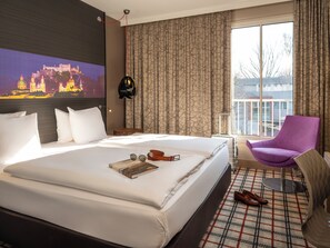 A hotel room with a large bed, a purple chair, a view of a cityscape, and a painting on the wall. - Mercure Salzburg City (Salzburg)