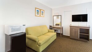 Ein Hotelzimmer mit gelber Couch, kleinem Kühlschrank, Schreibtisch mit Stuhl, Fernseher und Spiegel.