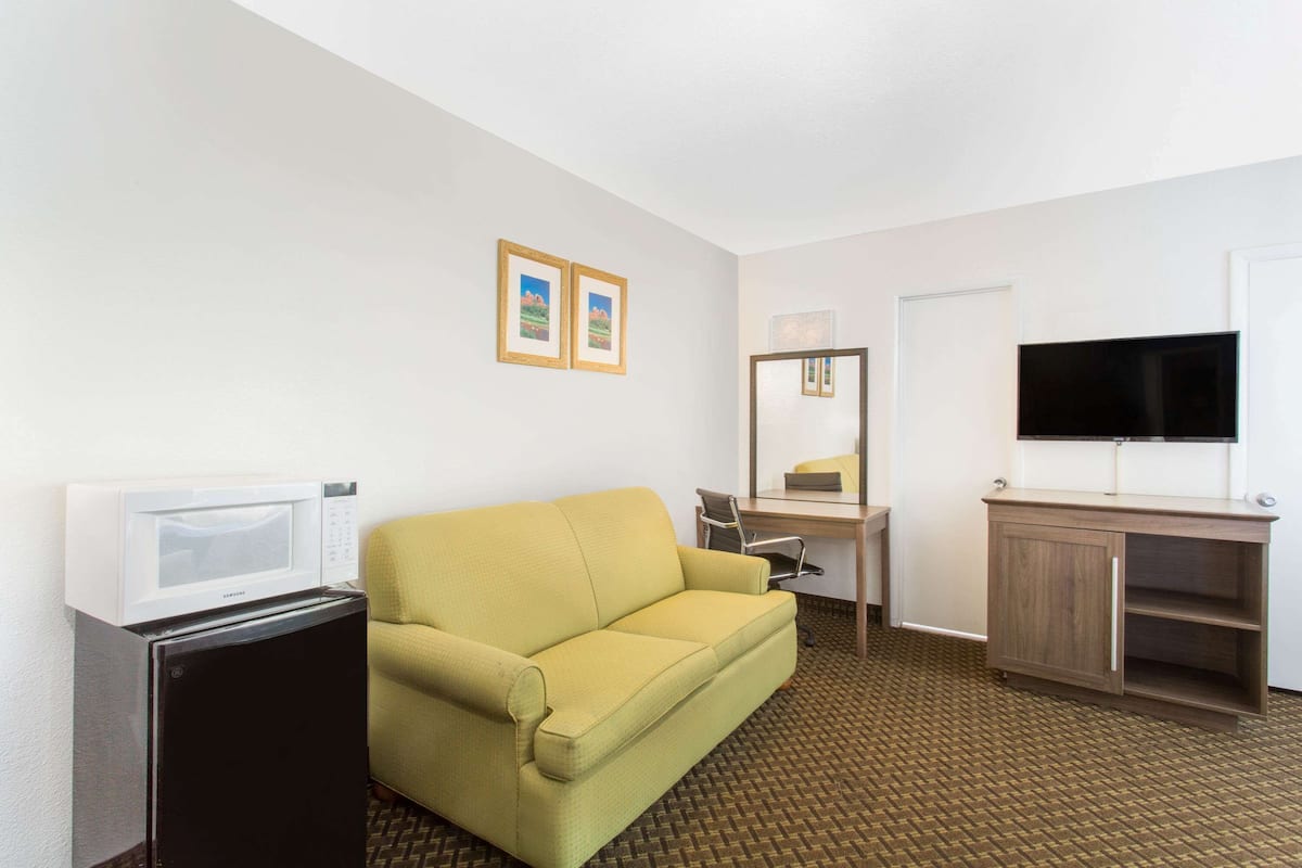 Ein Hotelzimmer mit gelber Couch, kleinem Kühlschrank, Schreibtisch mit Stuhl, Fernseher und Spiegel.