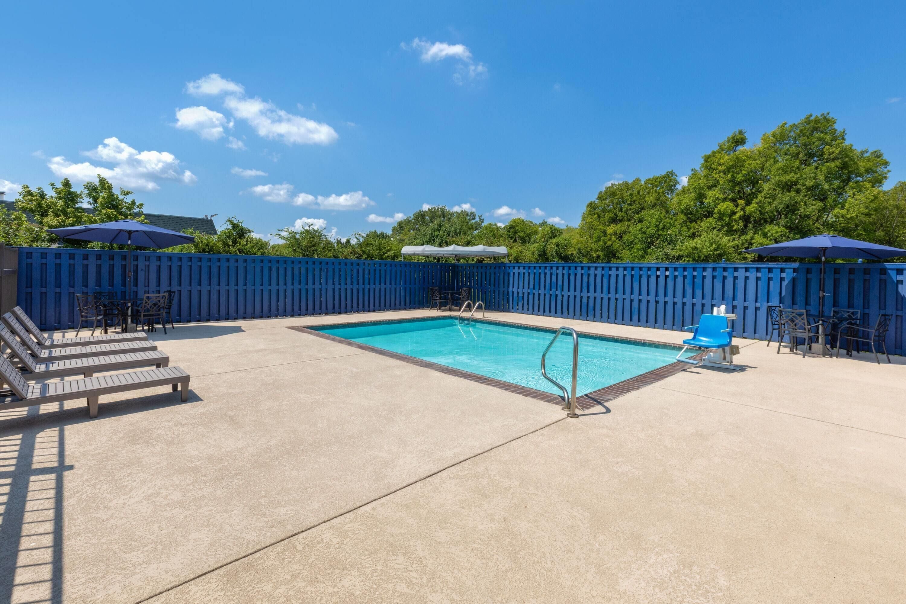 A rectangular swimming pool with a blue border, surrounded by a blue fence. There are lounge chairs and umbrellas around the pool.