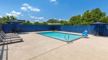 A rectangular swimming pool with a blue border, surrounded by a blue fence. There are lounge chairs and umbrellas around the pool.